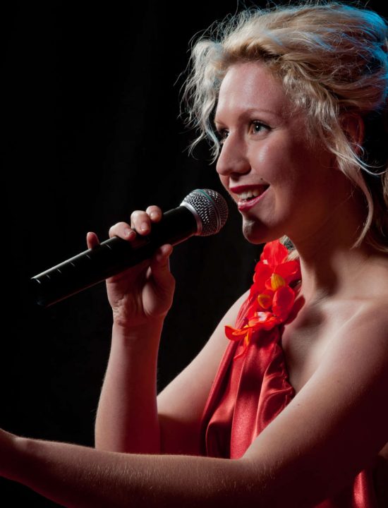 Sängerin im roten Kleid schaut lächelnd und singend ins Publikum und streckt den Arm nach vorne aus mit erhobenem Zeigefinger