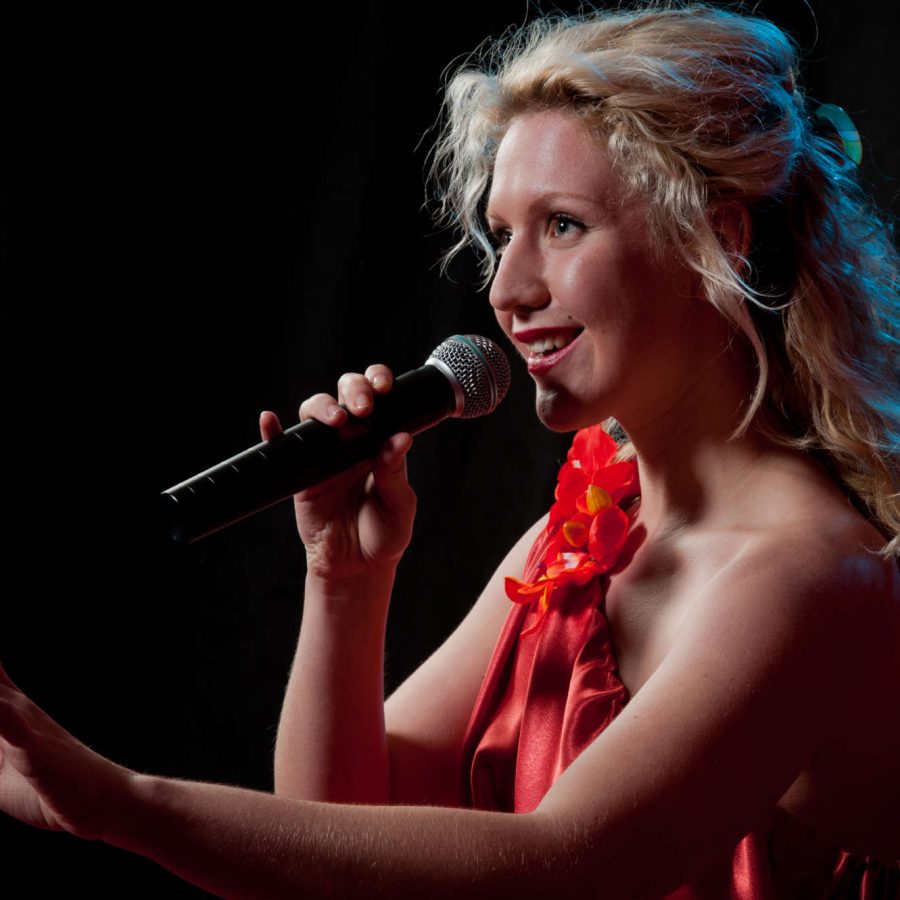 Sängerin im roten Kleid schaut lächelnd und singend ins Publikum und streckt den Arm nach vorne aus mit erhobenem Zeigefinger
