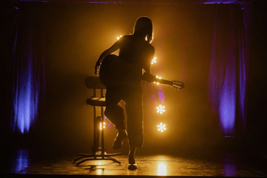 von hinten farbig beleuchtete Sängerin setzt sich mit der Gitarre auf einen Hocker auf der Bühne